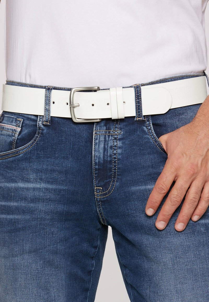 Weißer Leder-Gürtel mit einer Metallschnalle, getragen über blauen Jeans. Besitzt eine glatte Textur und mehrere Gürtellöcher für eine verstellbare Passform.