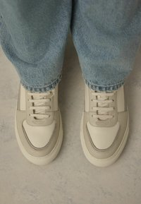 Helle beige Sneaker aus glattem Leder mit strukturierten Akzenten, mit Schnürdesign und Gummisohle, kombiniert mit verkürzten blauen Jeans.