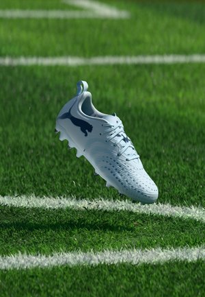 Chaussure de football blanche avec un logo bleu flottant au-dessus de l'herbe verte près d'une ligne blanche du terrain.
