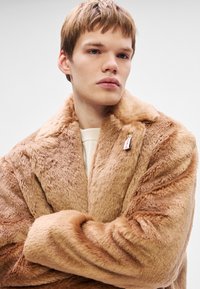 Sūnu kažoks gaismas brūnā krāsā ar mīkstu tekstūru, platā apkakle un neliels birkas piekabinājums. Modelis valkā krēmkrāsas džemperi zem tā.