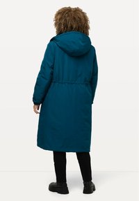 Tyrkysový péřový kabát s kapucí, elastickým pasem a dlouhými rukávy. Má hladkou texturu a sahá do poloviny lýtek. Nosit s černými kalhotami a botami.