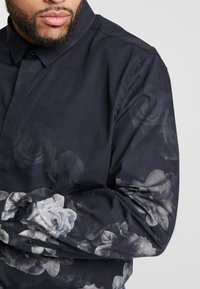 Homme portant une chemise boutonnée à manches longues sombre avec un motif floral gris subtil, bras croisés visibles, barbe et lèvres partiellement visibles.