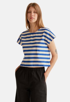 Joven con cabello liso castaño claro lleva una camiseta de manga corta a rayas azules y blancas y pantalones negros con las manos en los bolsillos.