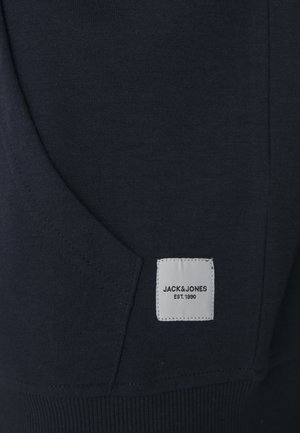 Navy blue sweatshirt material with a front pocket, ribbed hem, and a white label displaying "JACK & JONES EST. 1990." Textured fabric.