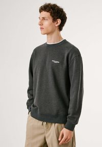 Sudadera gris de cuello redondo en tejido suave con puños y dobladillo acanalados. Presenta un pequeño logo blanco en el pecho. Textura suave, corte relajado.