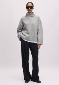 Grijze gebreide coltrui met geribbelde zoom, gecombineerd met zwarte wijde broek en zwarte vierkante schoenen. Model draagt een donkere zonnebril.