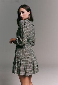 Robe à carreaux avec un col à revers, manches longues ballonnées et un ourlet évasé. Le tissu présente un motif beige et noir avec des accents bleus.