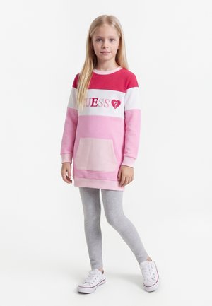 Młoda dziewczyna stoi, ubrana w długi różowo-czerwony sweter z logo "GUESS", szare legginsy oraz białe trampki na białym tle.