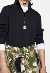 Personne portant un sweat-shirt noir à demi-fermeture éclair avec un logo blanc, des poignets de chemise blancs, et une veste à motifs verts, blancs et marrons nouée autour de la taille.