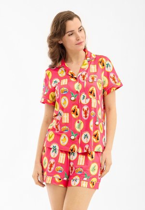 Mujer con conjunto de pijama de manga corta de color rosa brillante con patrones coloridos de frutas tropicales y langostas, de pie con una mano hacia abajo.