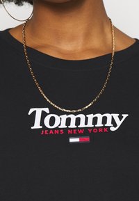 Svart T-shirt med texten "Tommy JEANS NEW YORK" i vitt och rött. Guldhalsband synligt. Mjuk material och rund halsringning.