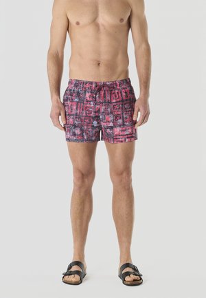 Hombre adulto sin camiseta, vistiendo pantalones de baño de patrón rojo y negro y sandalias negras, de pie contra un fondo claro y liso.