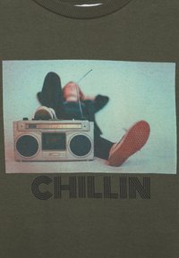 Oliwkowy sweatshirt z vintage'owym srebrnym boomboxem oraz grafiką osoby leżącej, z napisem "CHILLIN" wydrukowanym poniżej dużymi czarnymi literami.
