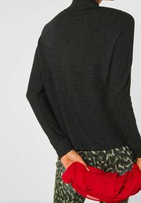 Suéter negro de cuello alto y puños acanalados. El modelo sostiene una prenda roja mientras lleva pantalones verdes con estampados.
