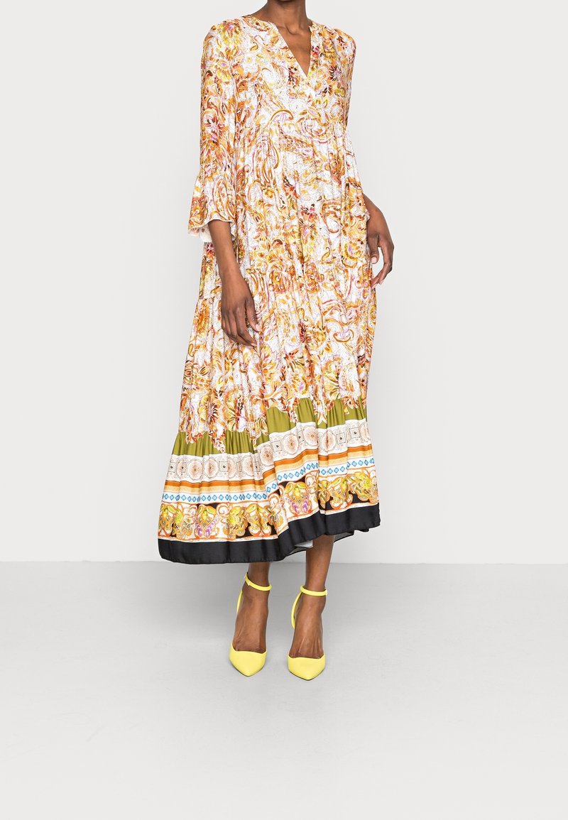 Nainen, jolla on yllään värikäs, kauniisti valuva maksimekko oransseilla, vihreillä ja mustilla yksityiskohdilla, yhdistettynä keltaisiin nilkkatuettuihin korkeisiin kenkiin, seisoo neutraalin taustan edessä.