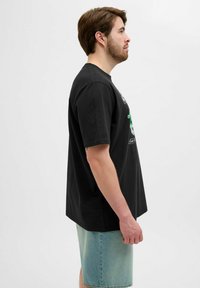Camiseta de algodón negra con mangas cortas, vista lateral. Presenta un estampado gráfico en verde y blanco. Corte holgado, combinada con pantalones cortos de denim azul claro.