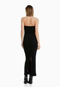 Robe longue noire sans bretelles à côtes, avec une silhouette ajustée et une fente haute sur le côté. Portée avec des bottines noires à plateforme.