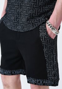 Shorts noirs avec un panneau latéral à motifs contrastants et une ceinture portant le texte "Project X Paris". Fabriqués dans un matériau lisse avec un design ajusté.