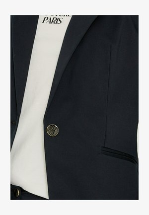 Blazer negro con botón geométrico dorado, usado sobre camisa blanca con texto "FUTURE PARIS".