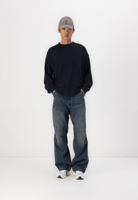 Ναυτικό μπλουζάκι με μακριά μανίκια και τσέπη, συνδυασμένο με φαρδιά τζιν παντελόνια σε ανοιχτό μπλε χρώμα με λεπτομέρειες patchwork, ολοκληρωμένο με λευκά αθλητικά παπούτσια και γκρι καπέλο.