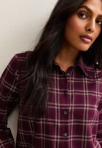 Chemise à carreaux bourgogne à boutons, fabriquée en tissu doux, dotée d'un col, de boutons noirs et de motifs à carreaux blancs, avec des manches longues.