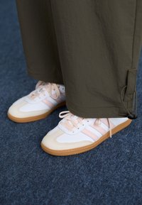 Baskets blanches avec des rayures roses et des semelles en caoutchouc, portées avec un pantalon olive sur un sol texturé bleu.