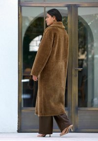Hnědý kožich z umělé kožešiny dlouhého střihu s vysokým límcem a volným střihem, doplněný hnědými kalhotami s širokými nohavicemi a světle barevnými podpatky. Hladká textura.