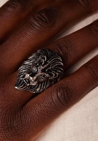 Zilveren ring in de vorm van een leeuwenkop op een donkerbruine hand, met gedetailleerde vachtstructuur en opvallende gelaatskenmerken, geplaatst tegen een lichte achtergrond.