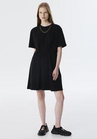 Schwarzes Kleid mit kurzen Ärmeln und taillierter Passform, mit Falten an der Taille. Ergänzt durch eine goldene Kettenhalskette und schwarze Sneakers.