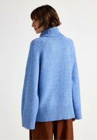 Kvinde med kort brunt hår iført en løs lyseblå turtleneck sweater og brune bukser, vendende mod venstre foran en ensfarvet baggrund.