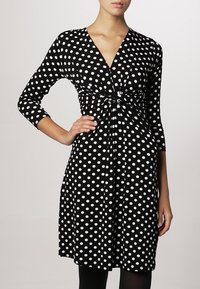 Svart klänning med vita prickar, V-iskärning, trekvartsärmar, rynkad midja och knälång kjol. Slätt tyg, figursydd design.