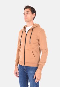 Felpa marrone con zip, cappuccio regolabile con coulisse, tasche frontali e dettagli neri sulla zip. Il tessuto è morbido e strutturato, indossato sopra una maglietta bianca.