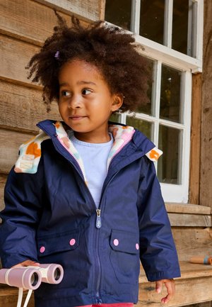 Jūras krāsas ūdensizturīga jaka ar rozā akcentu pogām, odere no flīsa daudzkrāsainā dizainā, valkāta virs gaiši zila krekla, turot binokļus.