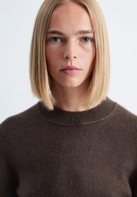 Blonde Frau mit glattem Haar, die einen braunen Rundhalsausschnitt-Pullover trägt, frontal vor einem schlichten hellen Hintergrund.