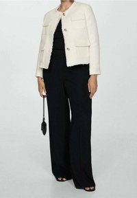 Witte textuurjacket met vier voorzakken en zilveren knopen, gecombineerd met een zwarte wijde broek en een zwarte handtas.