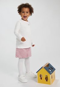 Witte lange mouwen shirt met de tekst "smile", roze denim rok, witte maillot en sneakers, naast een gele en blauwe houten vormen sorter.