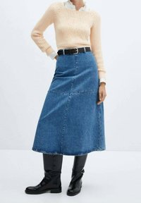 Jupe maxi en denim, silhouette trapèze, bleu clair, ourlet brut. Associée à un pull pêche texturé et des bottes noires montantes, soulignée par une ceinture noire.