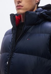 Colete acolchoado azul-marinho com gola alta e interior vermelho. Apresenta um fecho de correr frontal e um pequeno emblema perto da bainha. Tecido texturizado.