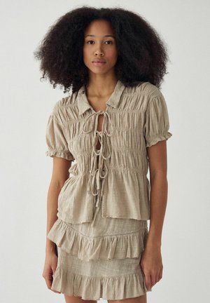 Kobieta o kręconych włosach, ubrana w beżowy marszczony top z zawiązywanymi sznureczkami oraz dopasowaną, warstwową spódnicę z falbankami, stojąca na jednolitym tle.