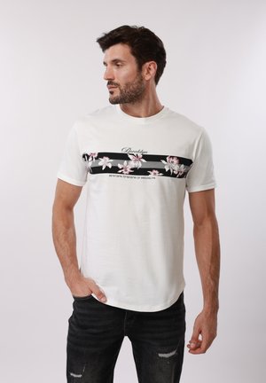 Άνδρας με γενειάδα που φοράει λευκό T-shirt με μαύρη λουλουδάτη ρίγα και το κείμενο "Brooklyn", σε συνδυασμό με μαύρο σκισμένο τζιν, στέκεται μπροστά σε απλό φόντο.