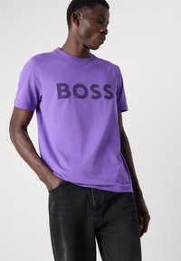 Fialové bavlněné tričko s velkým černým textem "BOSS" na přední straně, krátké rukávy, kulatý výstřih, v kombinaci s černými džíny.