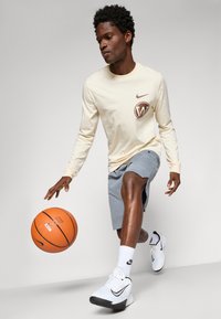 Pitkähihaista kerma väristä t-paitaa, jossa on ruskea Nike-logo, yhdistettynä harmaisiin shortseihin. Malli driblaa oranssia koriin pelaavaa palloa valkoisissa lenkkareissa.