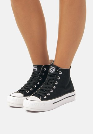 Zapatillas altas negras de lona, con suelas de goma blancas, cordones negros y un parche con el logo cerca del tobillo. Diseño clásico de punta redonda.