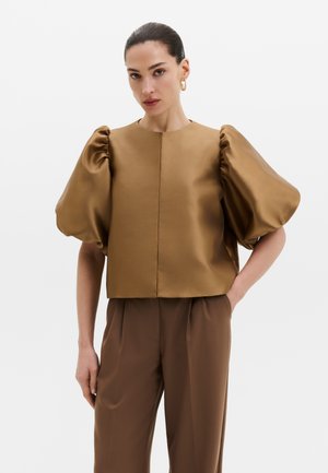 Femme aux cheveux foncés en chignon lisse, portant des boucles d’oreilles créoles dorées, un chemisier marron en satin à grandes manches bouffantes et un pantalon marron assorti.