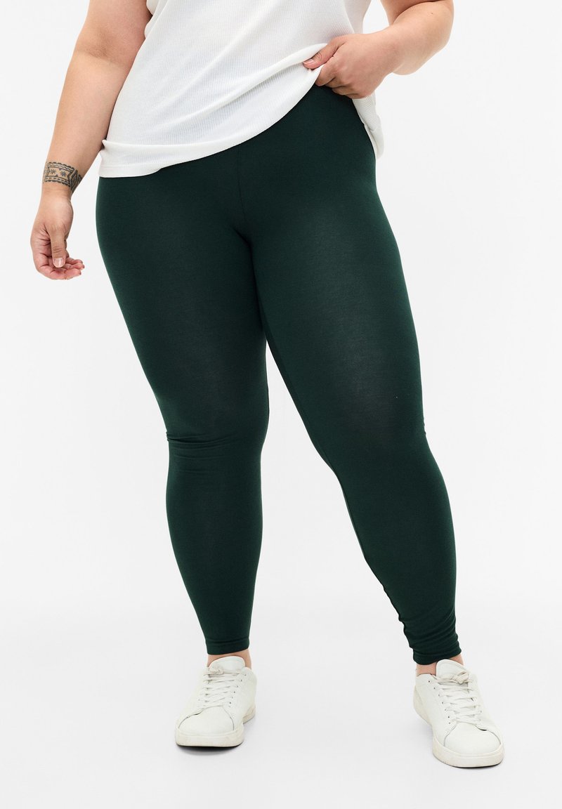 Groene leggings van rekbaar materiaal, met een gladde textuur en een aansluitend ontwerp. Gecombineerd met witte sneakers en een losse witte top.