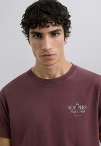 Camiseta de algodón color burdeos con mangas cortas, que presenta un logo blanco "The SCALPERS Taste & Talk" en la parte izquierda del pecho.