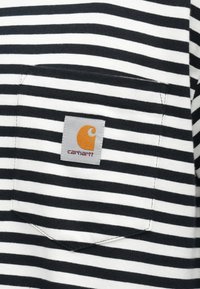 Μπλούζα με ασπρόμαυρες ρίγες και ένα γκρι ορθογώνιο μπαλώμα που φέρει πορτοκαλί λογότυπο. Η τσέπη βρίσκεται στο πάνω μέρος της πλάτης.