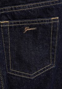 Jeans in denim blu scuro presentano una singola tasca posteriore con cuciture dorate a contrasto e un piccolo logo metallico "Guess".
