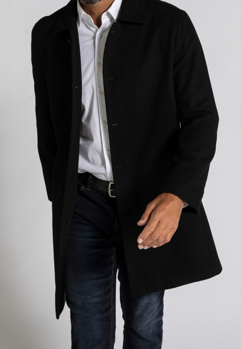 Cappotto di lana nera con frontale abbottonato, colletto e tasche laterali. Abbinato a una camicia bianca e jeans scuri, mette in risalto una silhouette slanciata.
