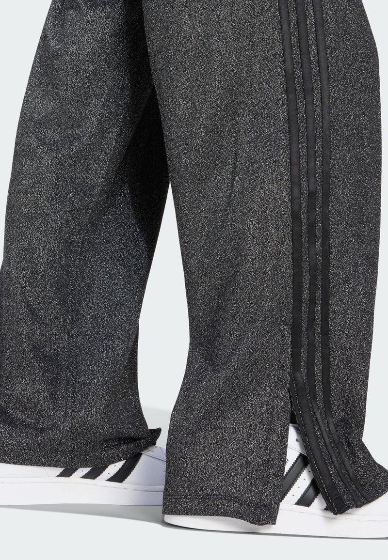 Pantalons de survêtement noirs pailletés avec des rayures noires sur les côtés et des fentes sur les côtés, associés à des baskets blanches avec des accents noirs.